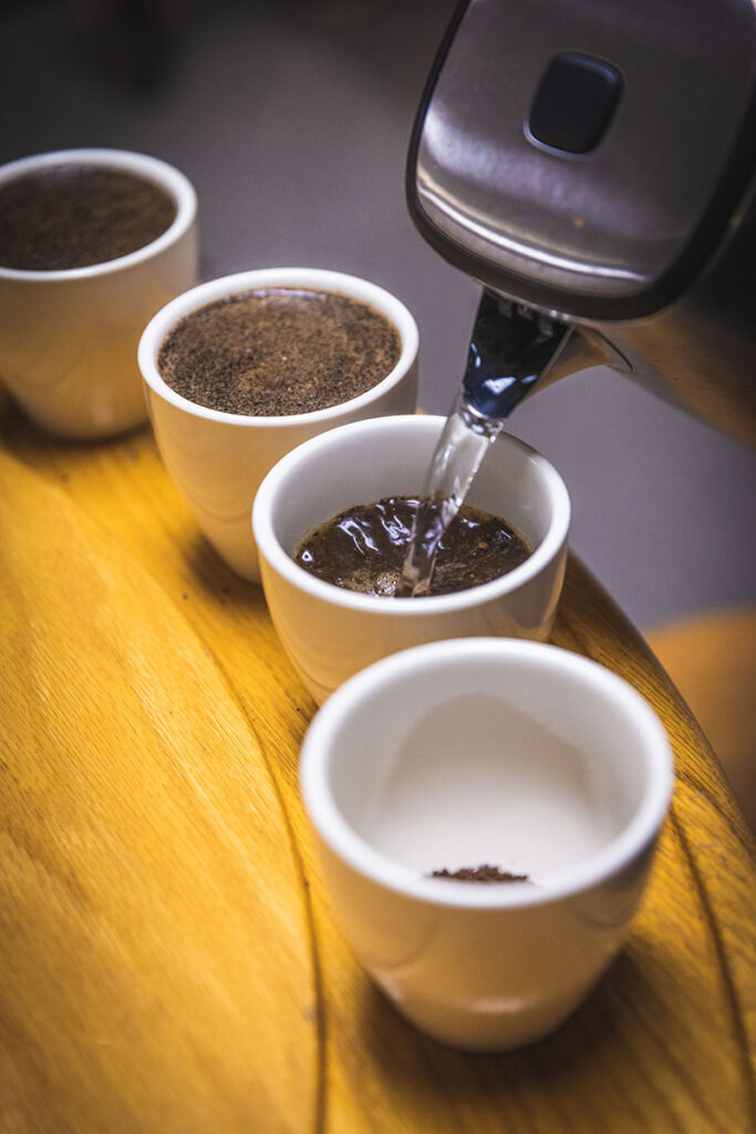 A kettle pouring hot water into a series of 4 cups during a coffee cupping