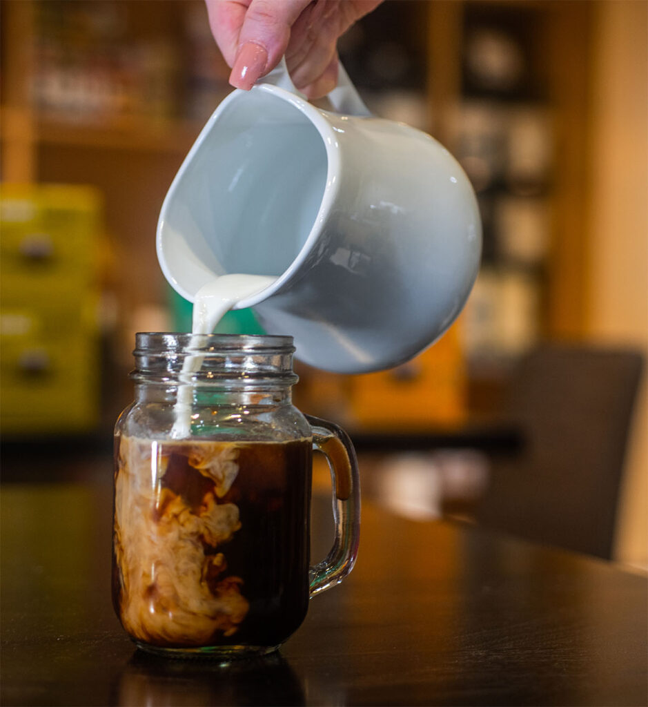A glass filled with iced coffee with milk being poured in from a white itcher