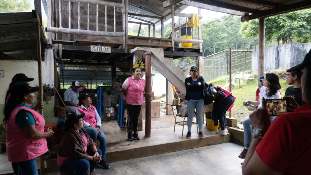 A group of women gathered to listen to a farmer talk about her coffee farm, open sided building, tropical setting.